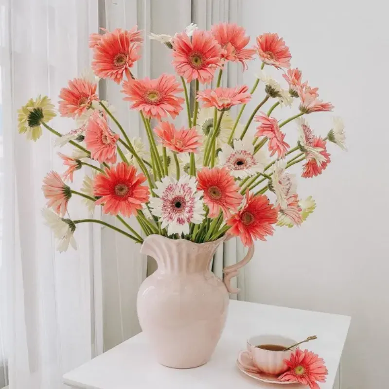 Bình hoa gerbera màu hồng và trắng trên bàn cùng cốc trà.